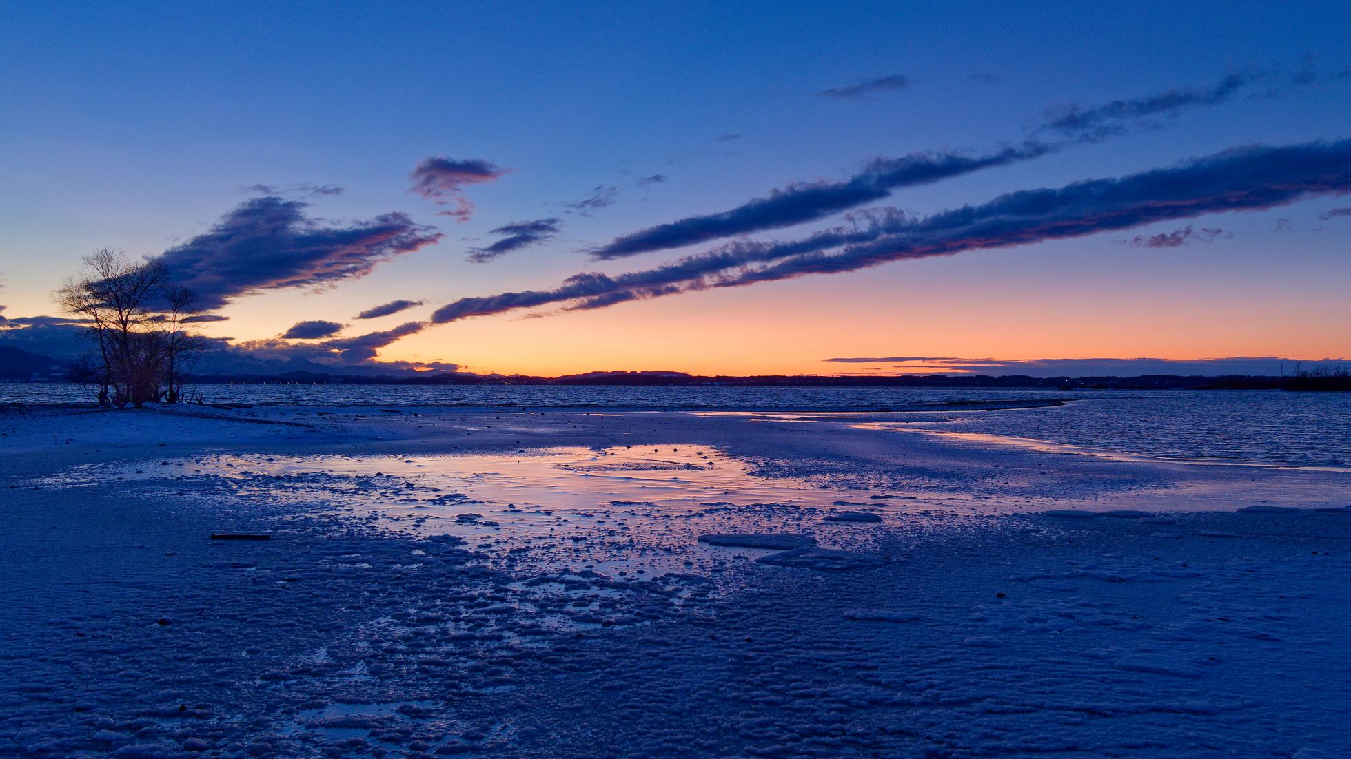 Blaue Stunde nach Sonnenuntergang an einem kalten Januarabend. Gefrorernes Uferwasser im Vordergrund am Horizont Berge und am dunkelrotblauen Himmel vereinzelten dunkle wolken.