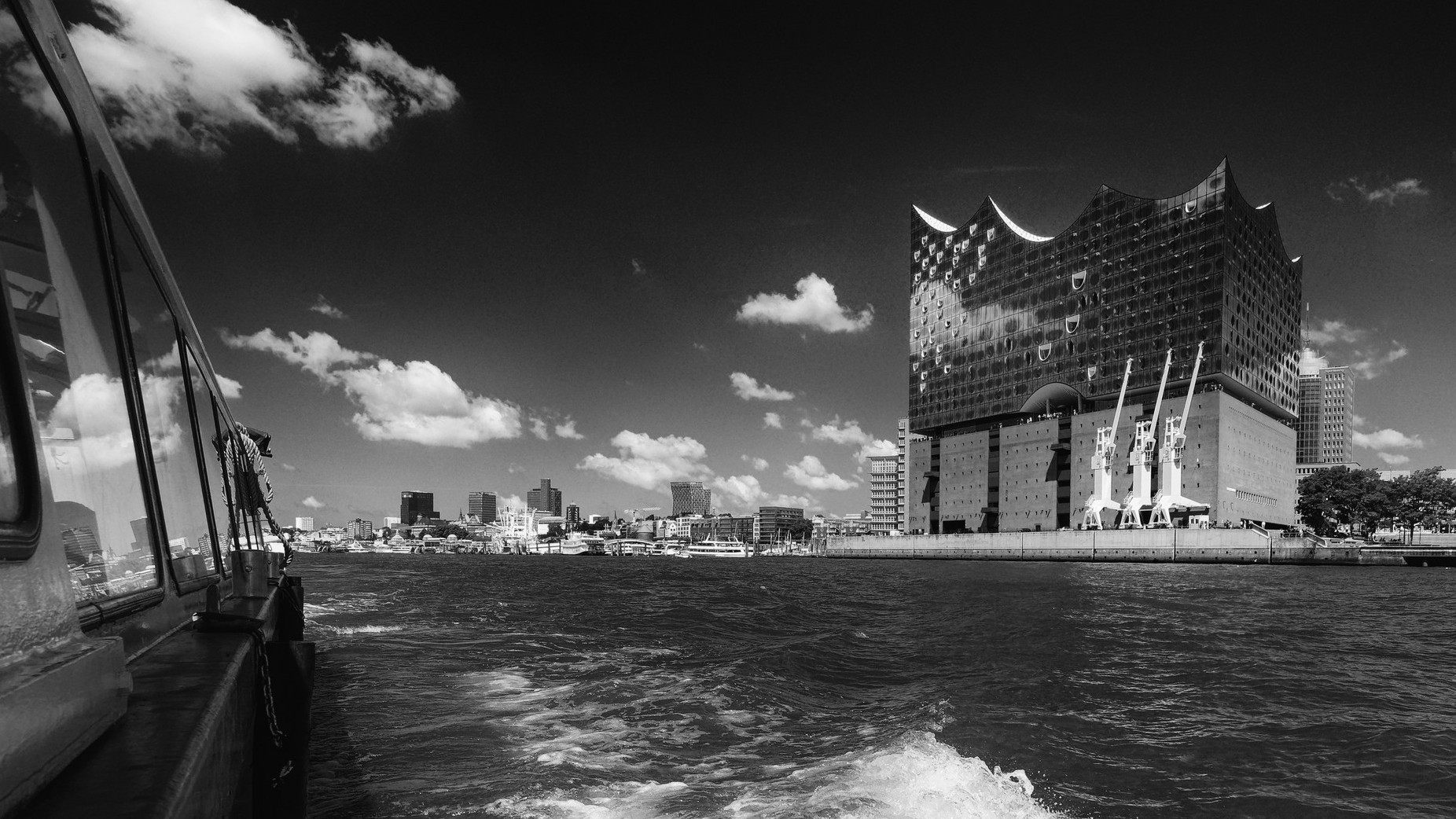 Blick über die Elbe von der Barkasse rückwärts auf die Landungsbrücken rechts die Elbphilharmonie. Fast schwarzer Himmel mit weißen Wolken.